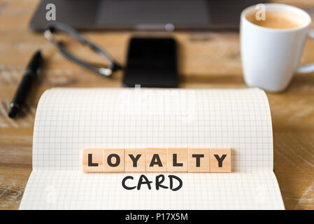 Closeup on notebook over vintage desk surface, front focus on wooden blocks with letters making Loyalty Card text. Stock Photo
