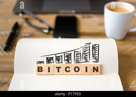 Closeup on notebook over vintage desk background, front focus on wooden blocks with letters making Bitcoin graph and rising arrow. Stock Photo