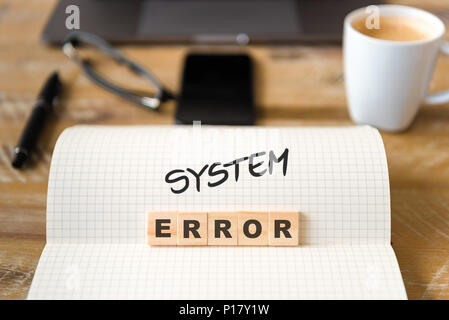 Closeup on notebook over vintage desk surface, front focus on wooden blocks with letters making System Error text. Stock Photo
