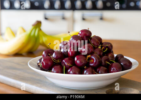 Close up of white ceramic bowl piled high with shiny, red cherries. Fruit bowl on UK kitchen table. Food, fruit photography. Stock Photo