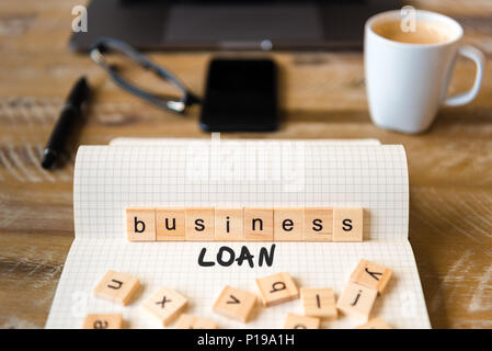 Closeup on notebook over vintage desk surface, front focus on wooden blocks with letters making Business Loan text. Stock Photo