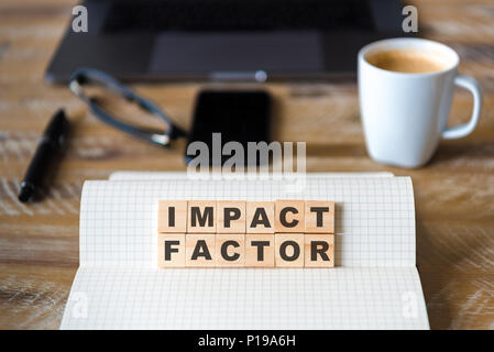 Closeup on notebook over vintage desk surface, front focus on wooden blocks with letters making Impact Factor text. Stock Photo