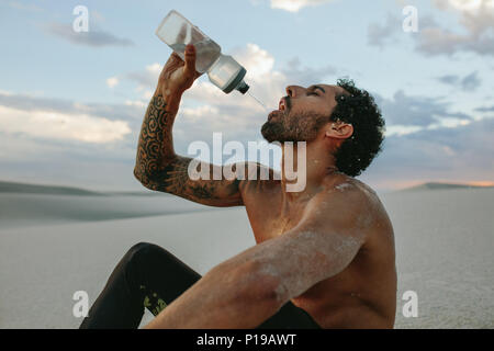 Athlete sitting on sand dune and drinking water. Man taking break after exercising in desert. Stock Photo