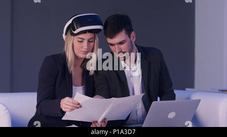 Female architect with VR headset showing blueprints to her male colleague using laptop Stock Photo