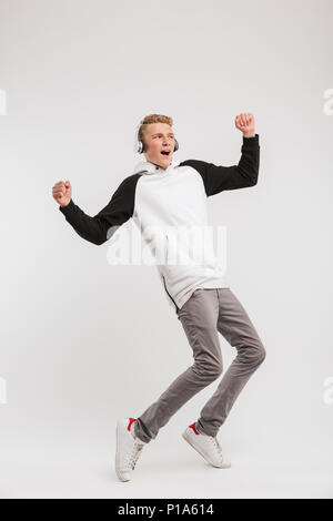 Full length portrait of teenage boy 16-18 years old wearing hoodie and wireless headphones having fun and dancing isolated over white background Stock Photo
