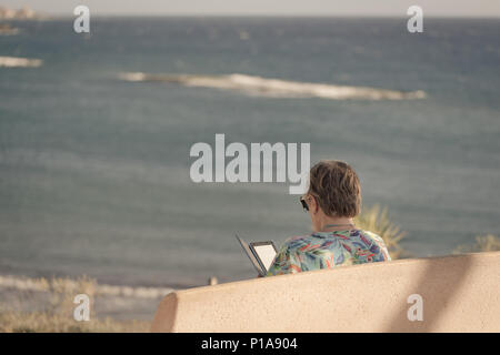 Older woman is sitting on bench with view on sea and she is reading ebook on her reader. Stock Photo