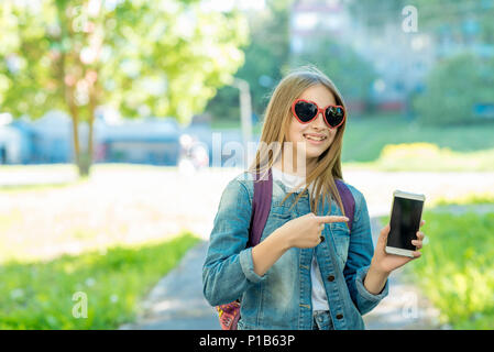 Little girl schoolgirl. In summer in park in fresh air. Holds hands of phone with a gesture of hand and point to the smartphone. Dressed in casual clothes in sunglasses in the form of hearts. Stock Photo