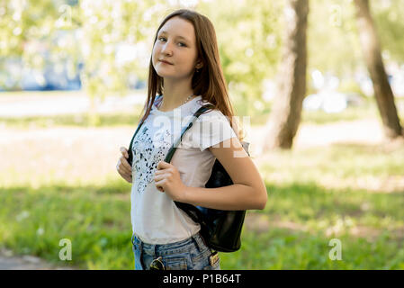 Beautiful teen girl in summer in a park outdoors in shade of a tree. Dressed in casual clothes. Behind the backpack. The schoolboy is going to school. Stock Photo