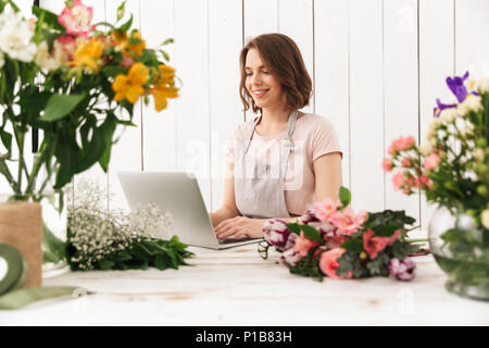 Young cute cheerful florist lady standing with flowers in workshop using laptop computer. Stock Photo