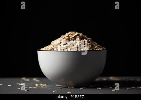 closeup of a white ceramic bowl full of rolled oats on a gray table, against a black background Stock Photo