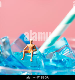 a miniature man wearing swimsuit and swim cap relaxing on the ice cubes of a blue cocktail served in a cocktail glass, against a pink background with  Stock Photo