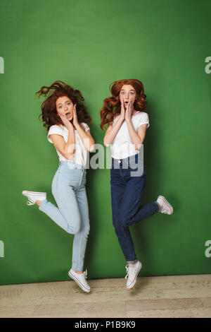 Full length photo of two surprised redhead girls 20s in white t-shirts and jeans touching cheeks with raised legs isolated over green background