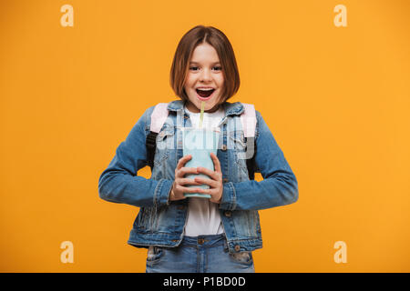 Portrait of a cheerful little schoolgirl with backpack holding plastic cup over yellow background Stock Photo