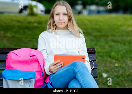 Little girl blond schoolgirl. Summer in nature. Sits on bench in the hands holding a tablet. After classes, she relaxes listening to music. Enjoys relaxation after school. Stock Photo