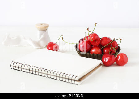Feminine still life composition. Sweet red cherry fruit in little metal bowl, blank notebook and spool of silk ribbon on white table. Defocused background. Summer rustic scene. Styled stock photo. Stock Photo