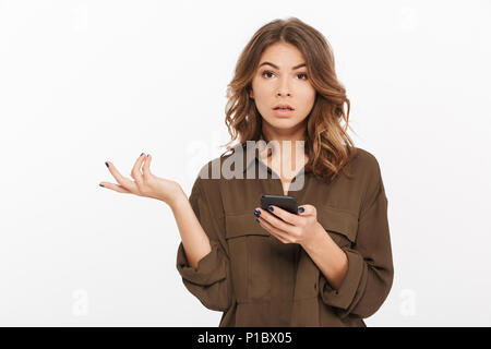 Portrait of a confused young woman holding mobile phone and looking at camera isolated over white background Stock Photo