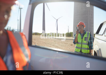 Smiling engineer using walkie-talkie at truck at sunny wind turbine power plant Stock Photo
