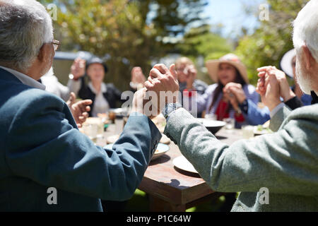 Active senior friends holding hands, praying at sunny garden party table Stock Photo