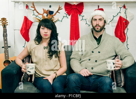 A couple (20's-30's) (twenties-thirties) holding mugs and sitting on a sofa posting for an ironic holiday portrait. Stock Photo