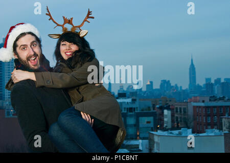 A couple standing on a brooklyn rooftop with the manhattan skyline behind them, posing for a christmas portrait. Stock Photo