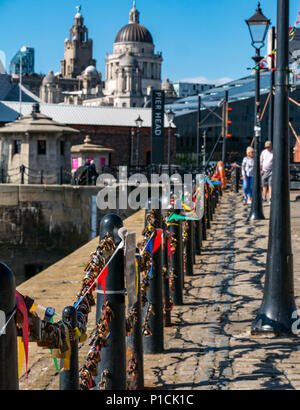 Albert Dock, Liverpool, England, United Kingdom, 11th June 2018. UK weather: sunshine on the Mersey. A beautiful sunny day with blue sky along the River Mersey in Liverpool today for visitors and locals. Railings are covered with love locks on the promenade. The domes of Port of the Liverpool building and towers of the Royal Liver building are in the distance. A senior couple walk along the promenade Stock Photo