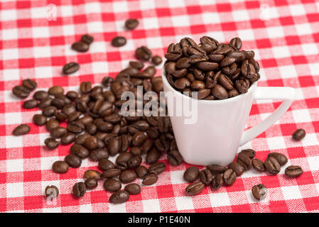 A white coffee mug is  filled with brown coffee beans. The coffee beans are also spread out on the kitchen table with a red checkered tablecloth as a  Stock Photo