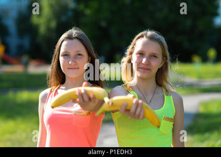 Two girl friends. Summer in nature. Happy extends a hand in the hands of banana. Healthy lifestyle. Proper nutrition of children. Useful fruit for school children. Stock Photo