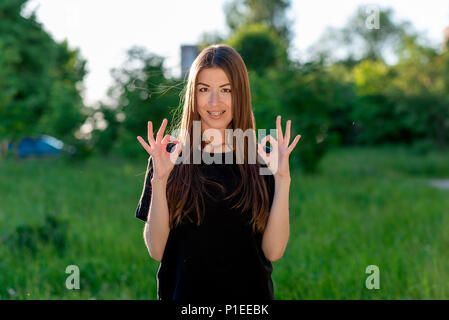 Summer in the fresh air young beautiful brunette asian girl. Two hands showing OK. Emotional positive attitude. Happy smiling teeth white braces. Stock Photo