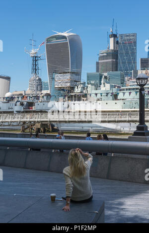 a young woman with blond hair sitting on a wall at the side of the river thames looking at the city of London skyline in the distance. attractive girl Stock Photo