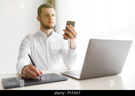 Young smiling bearded businessman is working on computer holding smartphone. Stock Photo