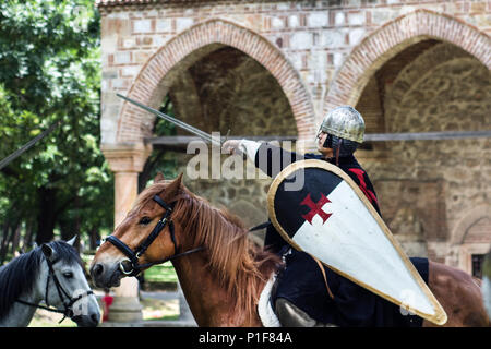 Nis, Serbia - June 10, 2018: Medieval knights posing on brown horse with armor, shield and sword in air and old building in background. Middle ages Stock Photo