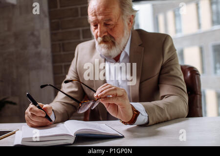 old exhausted man is putting his glasses on the desk while making notes Stock Photo