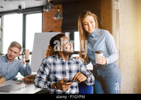 smiling Afro guy in checked T-shirt is looking at shy office worker holding a cup of tea Stock Photo