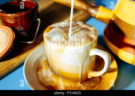 milk being poured into an overflowing mug of iced coffee on the table with old fashioned 70s kitchenware Stock Photo