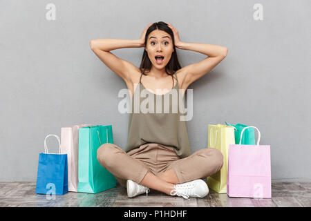 Full length portrait of a shocked young asian woman sitting on a floor with shopping bags over gray background Stock Photo