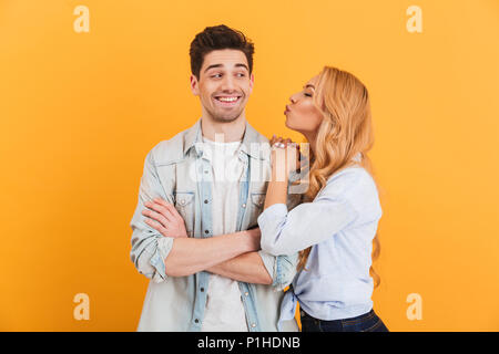 Portrait of young lovely people in basic clothing expressing love and affection while woman kissing man on cheek isolated over yellow background Stock Photo
