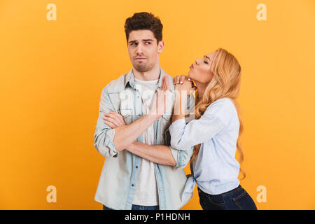 Portrait of young displeased man gesturing to stop with hand while beautiful woman kissing his cheek isolated over yellow background Stock Photo