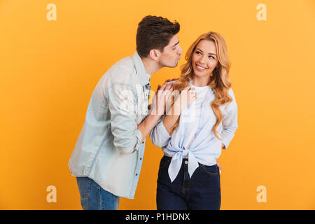 Image of young people wearing denim clothing in relationship expressing love and affection while man kissing woman on cheek isolated over yellow backg Stock Photo