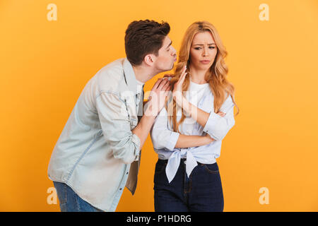 Portrait of outraged displeased woman gesturing to stop with hand while man trying to kiss her isolated over yellow background Stock Photo