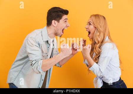 Portrait of joyful young couple man and woman smiling and clenching fists while looking at each other isolated over yellow background Stock Photo