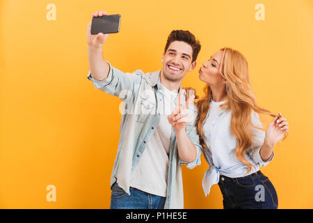 Portrait of lovely couple taking selfie photo on cell phone while woman kissing man on his cheek isolated over yellow background Stock Photo