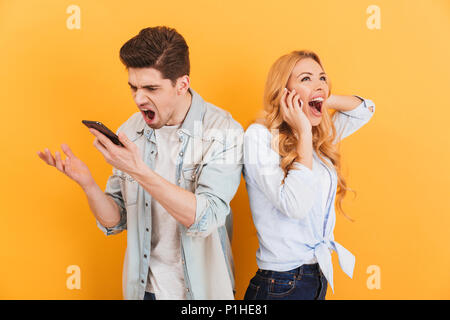 Image of furious man screaming in smartphone while happy woman having pleasant mobile conversation isolated over yellow background Stock Photo