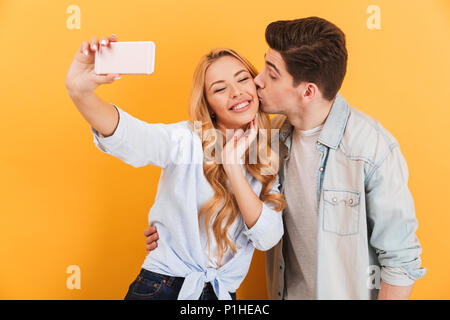 Portrait of lovely couple taking selfie photo on mobile phone while man kissing woman on cheek isolated over yellow background Stock Photo