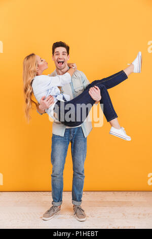 Full length photo of cheerful man smiling and holding woman in his arms who kissing his cheek isolated over yellow background Stock Photo