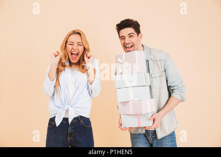 Image of surprised woman shouting in happiness while handsome man holding lots of gift boxes isolated over beige background Stock Photo