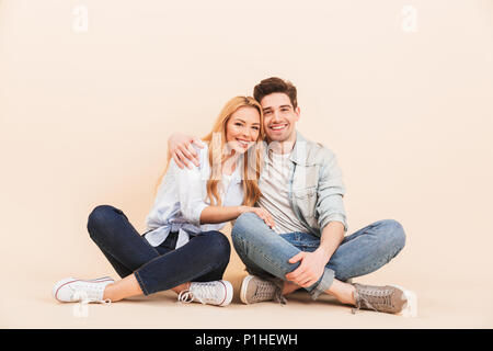 Image of nice and cute couple in denim clothing smiling and hugging together while sitting on the floor with legs crossed isolated over beige backgrou Stock Photo