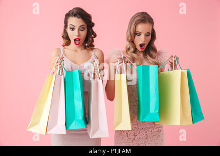 Two shocked women in dresses opening their packages with purchases over pink background Stock Photo