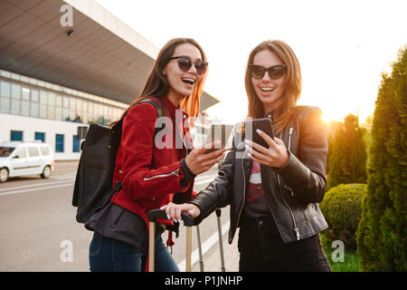 Photo of two beautiful european women wearing sunglasses using mobile phones while standing with luggage near airport waiting for flight or after depa Stock Photo