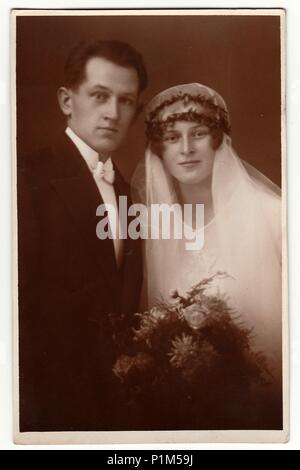 THE CZECHOSLOVAK  REPUBLIC - CIRCA 1930s: Vintage photo of newlyweds. Bride wears a long veil and holds wedding bouquet. Groom wears black suit and white bow tie. Black & white antique studio portrait. Stock Photo