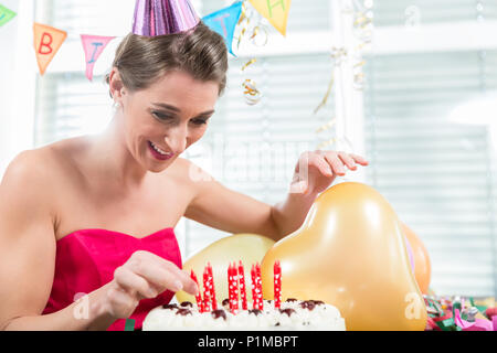 Portrait of a beautiful woman smiling while putting red candles on a birthday cake for her anniversary indoors in a decorated room Stock Photo